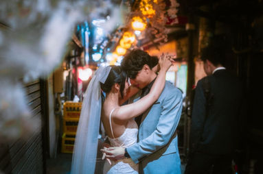 A bride and groom share an intimate moment as they dance together in a narrow Tokyo alley, illuminated by soft, colorful lights from lanterns. The bride, wearing a wedding dress and veil, is embraced by the groom, who is dressed in a light blue suit. In the background, a man walks by, adding a dynamic, city vibe to the romantic scene.