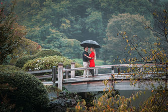 A couple stands close beneath a single black umbrella on a rain-washed bridge inside a quiet Japanese garden. The soft mist and lush greenery wrap around them, turning the embrace into something tender and almost suspended in time.