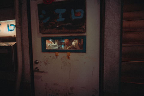 A worn metal door with a small rectangular window reveals people inside a dimly lit Tokyo bar or kitchen. Peeling paint and handwritten Japanese text emphasize the raw, everyday texture of the city at night. Photograph by Deniz Demir in Tokyo.