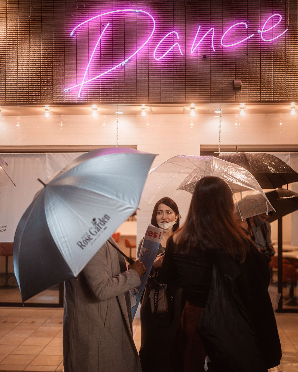 Thumbnail: A group of people standing under umbrellas outside a neon-lit "Dance" sign at night.