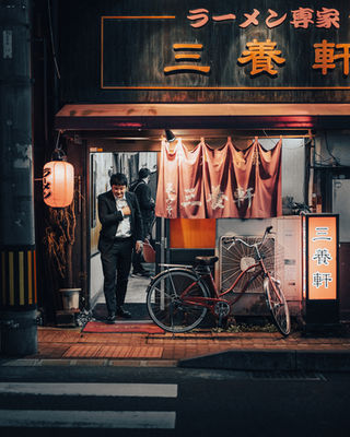  A man walks out of a ramen restaurant in Tokyo, passing under the red hanging curtains and next to a vintage bicycle. The warm lighting highlights the inviting and authentic ambiance of the ramen shop. NFT Photography by Tokyo Photographer Deniz Demir. Tokyo, Japan.