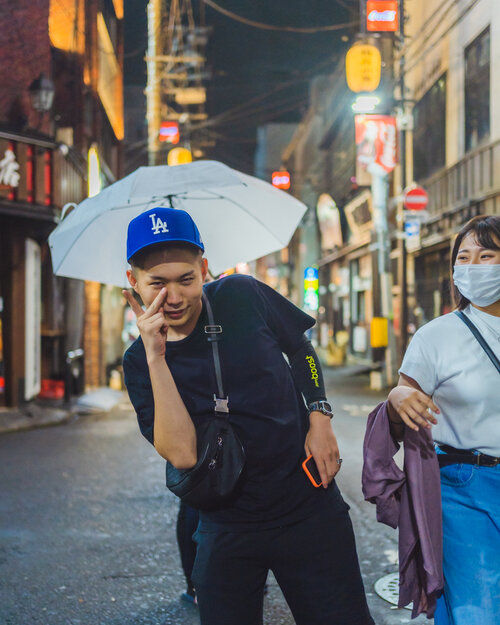 A young man in a blue cap playfully poses on a rainy Tokyo street at night, holding an umbrella, while a masked woman walks beside him under city lights.