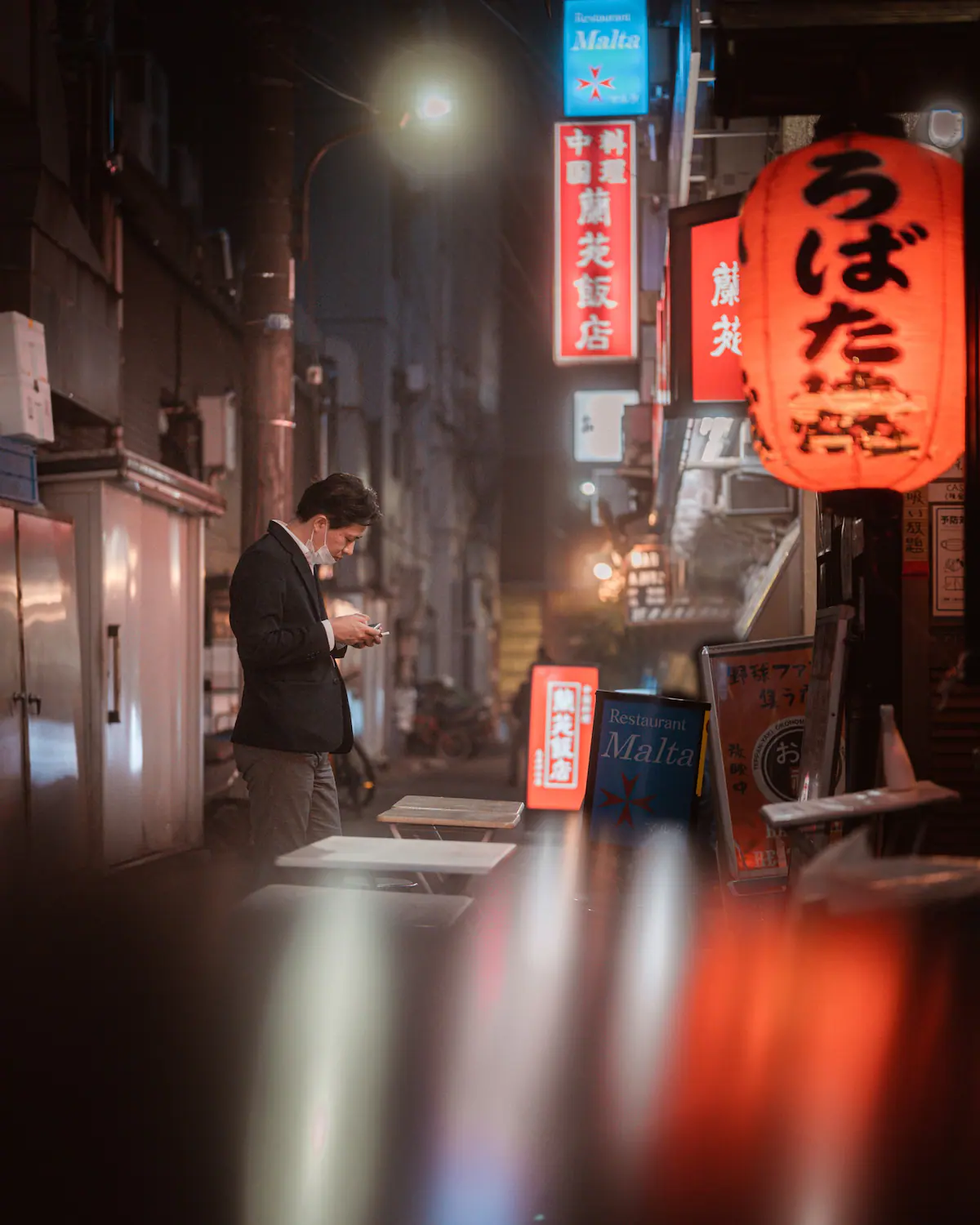  A businessman in a dark suit stands in a narrow Tokyo alleyway at night, illuminated by neon signs and lanterns, lost in thought.