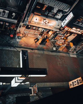 This bird's-eye view captures a busy street in Tokyo with bustling pedestrians walking past a restaurant. The vibrant signage and patterns of the street contrast beautifully against the calm of the evening. NFT Photography by Tokyo Photographer Deniz Demir. Tokyo, Japan.