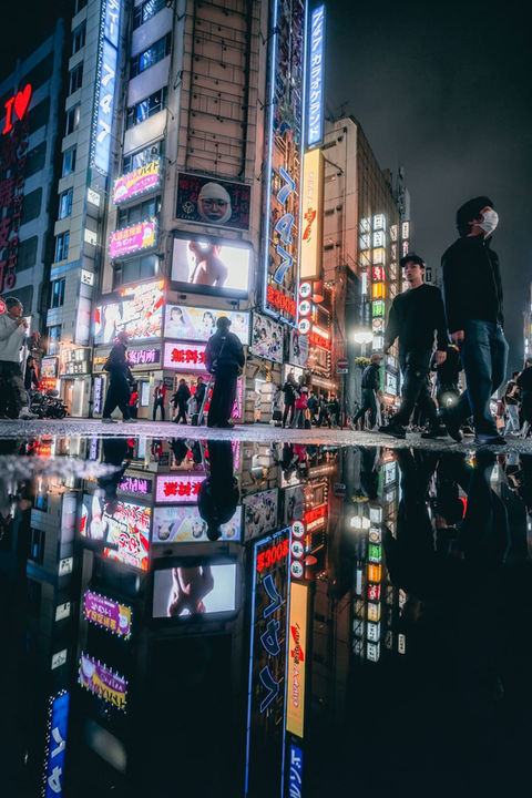 Pedestrians cross a neon-lit street in Shinjuku at night, reflected sharply in a large puddle on the pavement. Bright signs for karaoke, bars, and entertainment venues tower overhead, while masked figures move through the frame, blending motion, reflection, and light. Night street photograph taken in Tokyo by Deniz Demir.
