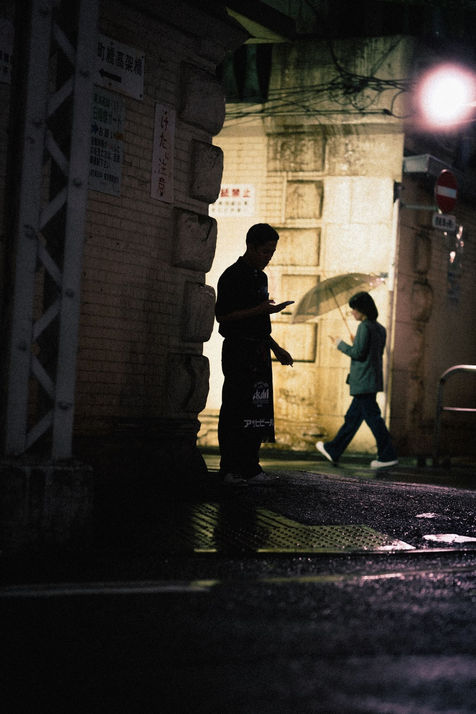 A dimly lit street scene features a person standing with a phone, while another walks in the distance under an umbrella.