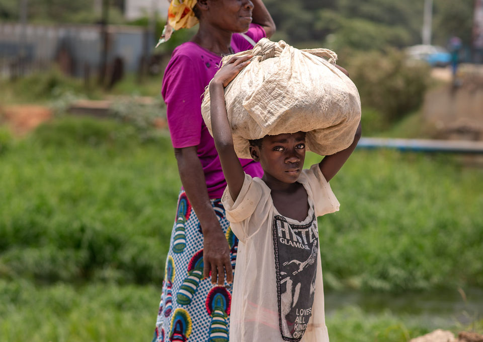 A woman and a child in traditional dress walking outdoors while carrying a woven basket balanced on her head.