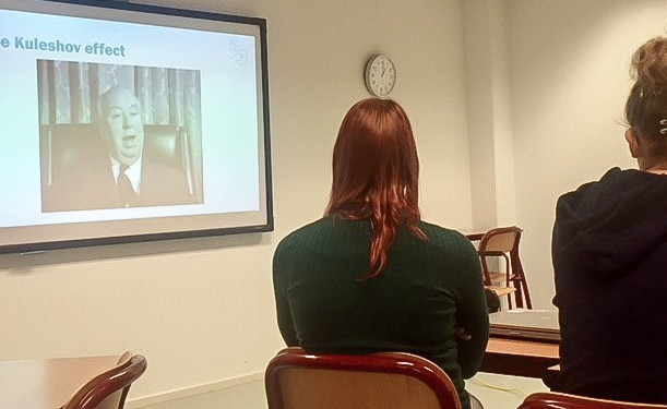 Foto van twee studenten die naar een projectiescherm zitten te kijken. Op het projectiescherm is een foto van het Kuleshov effect te zien.