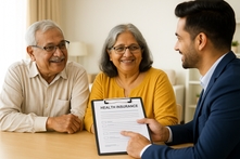 Elderly couple smiling as a man in a blue suit discusses a health insurance document in a bright office. The mood is reassuring and positive.