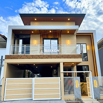 Modern two-story furnished house with a sleek contemporary design, featuring warm wood accents, glass balcony railings, ambient exterior lighting, and a gated carport for two vehicles under a bright blue sky. The facade shows clean architectural lines and the logo “Brixon Realty International” at the center.
