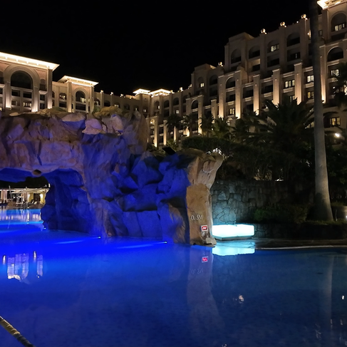 夜のホテルの屋外プール。青く照らされた水面と岩の洞窟風装飾、背後にライトアップされた高層建物。岩に0.5M表示