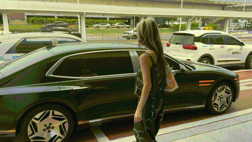 空港の車寄せで、黒いセダンの横に立つ女性。白いタクシーが並び、明るい昼の雰囲気。