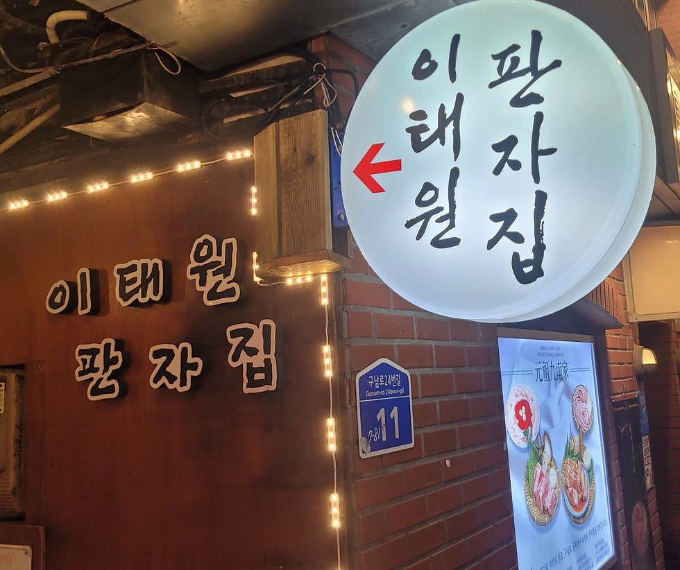 韓国料理店の外観。丸い看板に韓国語の店名と赤い矢印、木壁に店名文字、料理写真の案内板が見える。