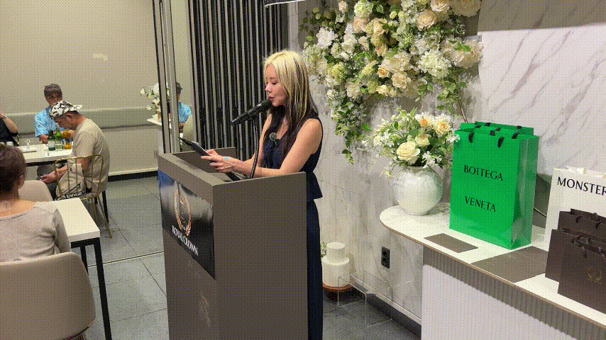 室内のイベント会場で、金髪の女性が演台でマイクに向かって話し、横に白い花と緑のBottega Venetaの袋が並ぶ。