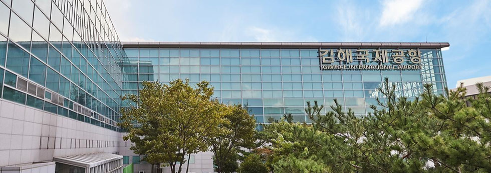 ガラス張りの金海国際空港の建物。上部に「GIMHAE INTERNATIONAL AIRPORT」の看板があり、手前に木々、青空が広がる。
