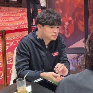 カジノ会場で、黒いジャケットの男性が女性相手にチップを扱う。赤い柄の床とロープ柵があり、胸にNO GAMBLING NO FUTUREの文字が見える。