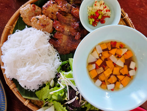 竹ざるの上に、焼き肉、米粉の麺、野菜、ニンジンとパプリカ入りのスープ、刻んだチリとニンニクがある。色鮮やかなベトナム料理。