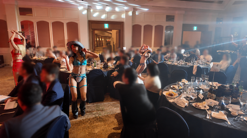 宴会場で、赤や青の衣装と帽子の女性ダンサーがテーブル間で踊り、着席した客が見守る。温かな照明と料理皿が並ぶ。