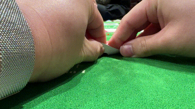 緑のフェルトの上で、両手が小さな白い駒をつまんでいる接写。左に銀色の袖が見え、静かな集中感がある。