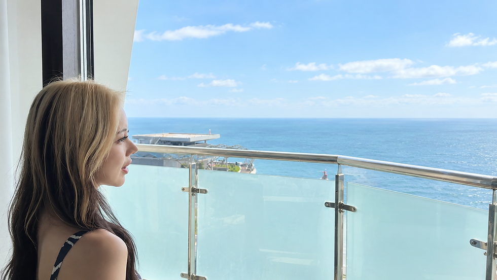 海沿いのバルコニーで、女性が青い海と空を静かに見つめている。ガラス手すり越しに明るい景色が広がる。