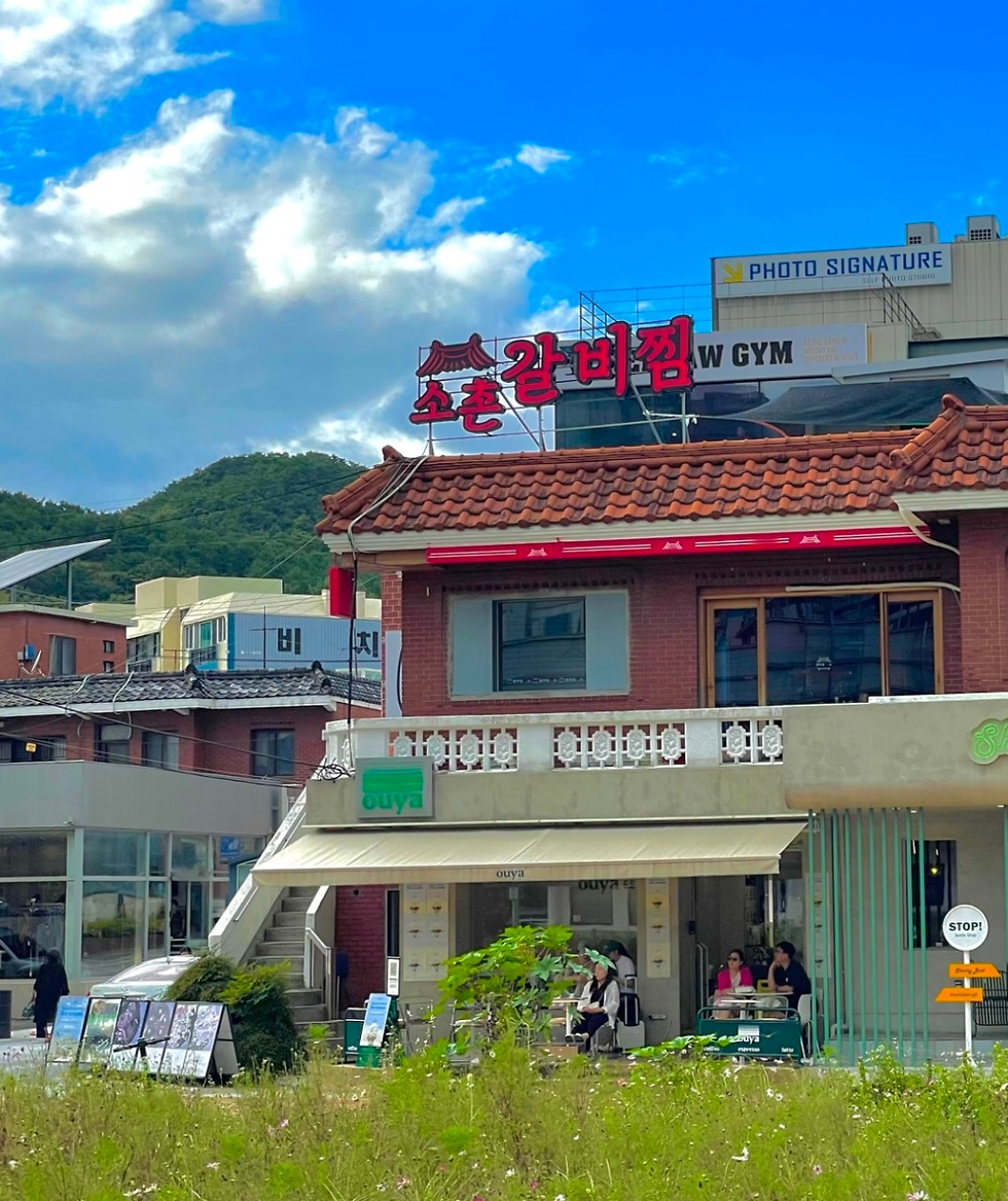 青空と雲の下、韓国語看板「소촌갈비찜」が載る建物とカフェ「ouya」の前で人々が座る、のどかな街並み。