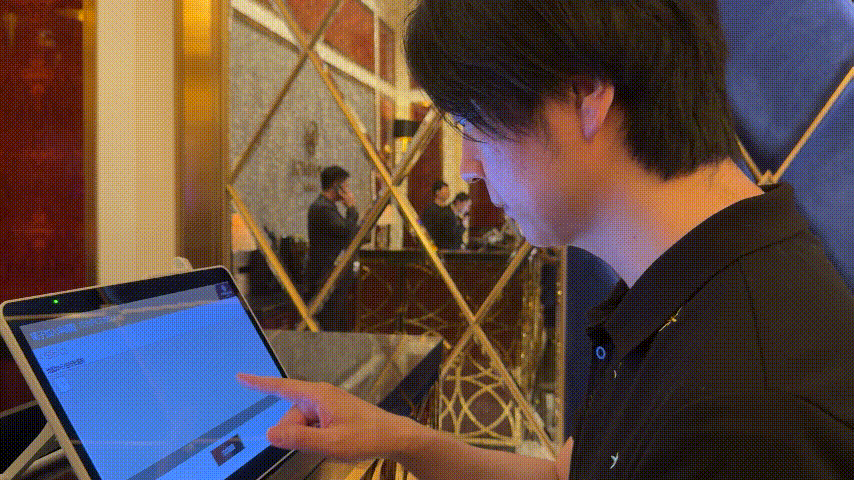 暖色の室内で、黒い服の男性がタブレット画面を指で操作している。鏡張りの背景に人影が映り、集中した雰囲気。
