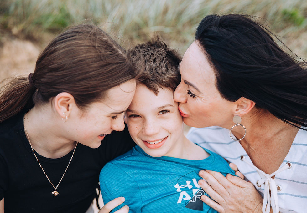 A little girl on the left side of the photo is happily facing her little brother in the middle while their mother kisses him on the cheek on the right.