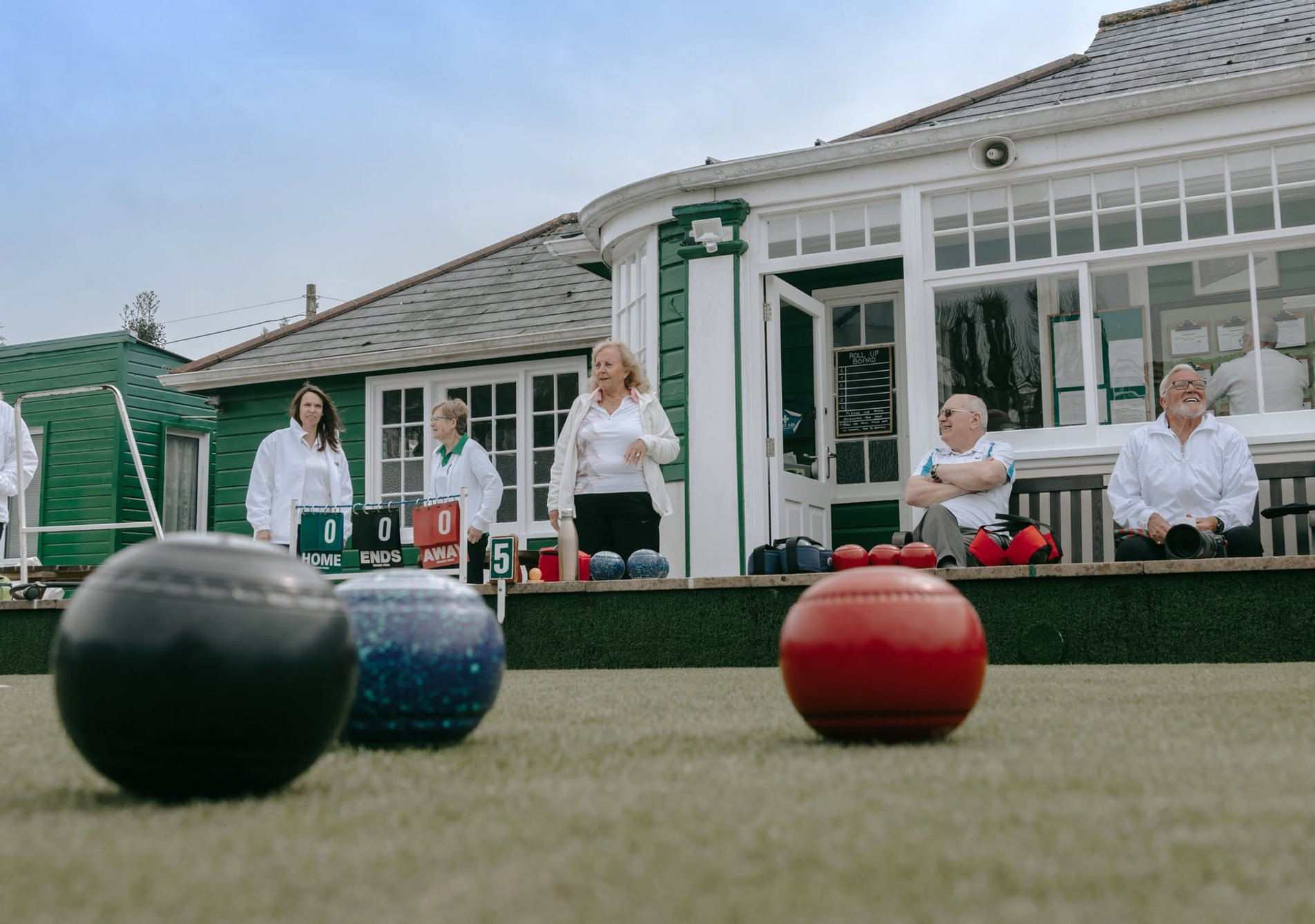 Lawn bowls players in white stand by a green clubhouse, with colorful bowls in the foreground and a scoreboard showing 0-0.