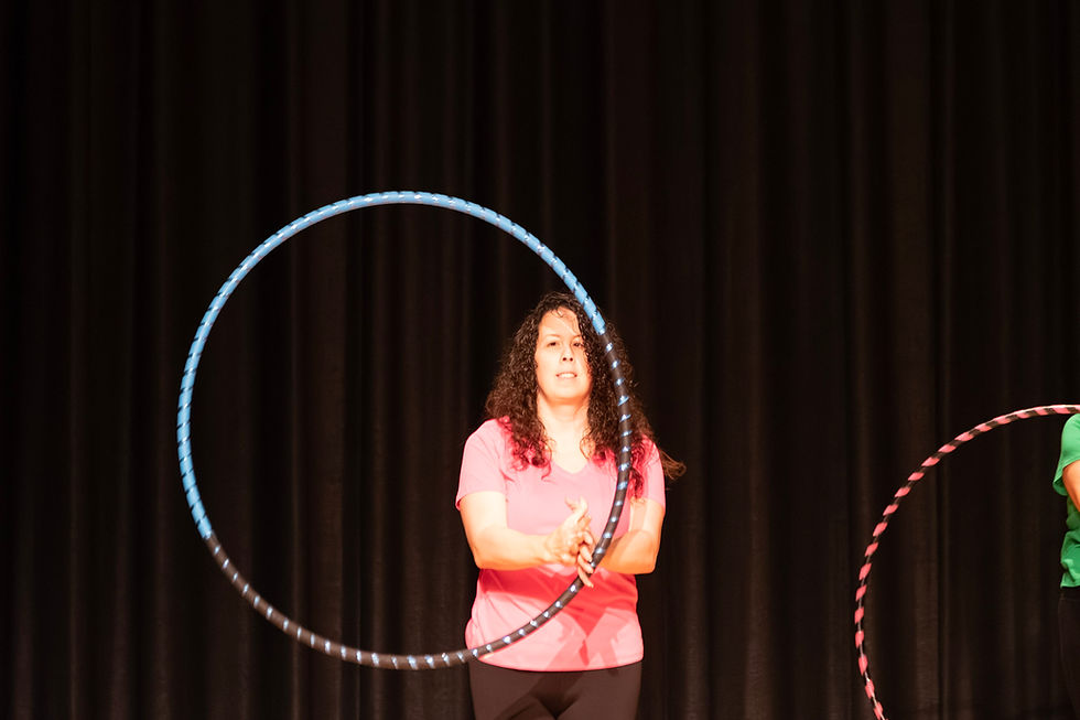 Andi is wearing a light pink top and black pants. She is using a blue and black hula hoop, and it is currently rotating in the air in front of her as she hand hoops. There are black stage curtains behind her.