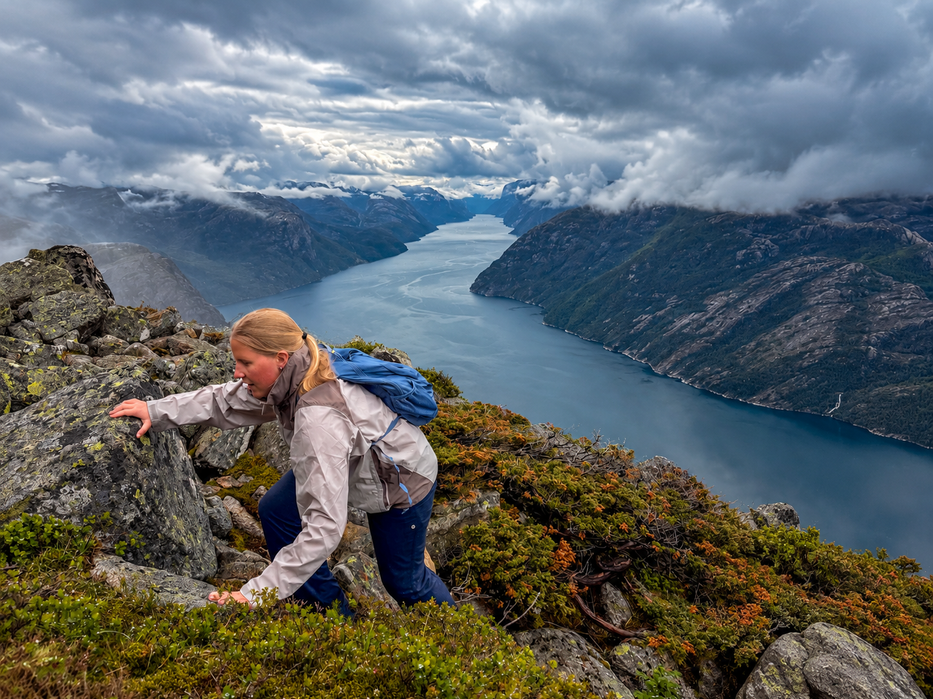 Hiker scrambling up rocky terrain with sweeping fjord views under dramatic clouds.
