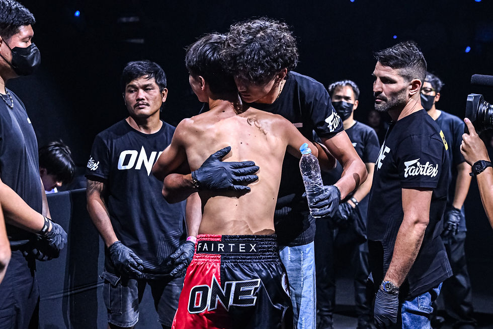 Boxer in red "ONE" shorts gets a supportive hug backstage from a team member, flanked by others in "ONE" shirts. Mood is emotional.