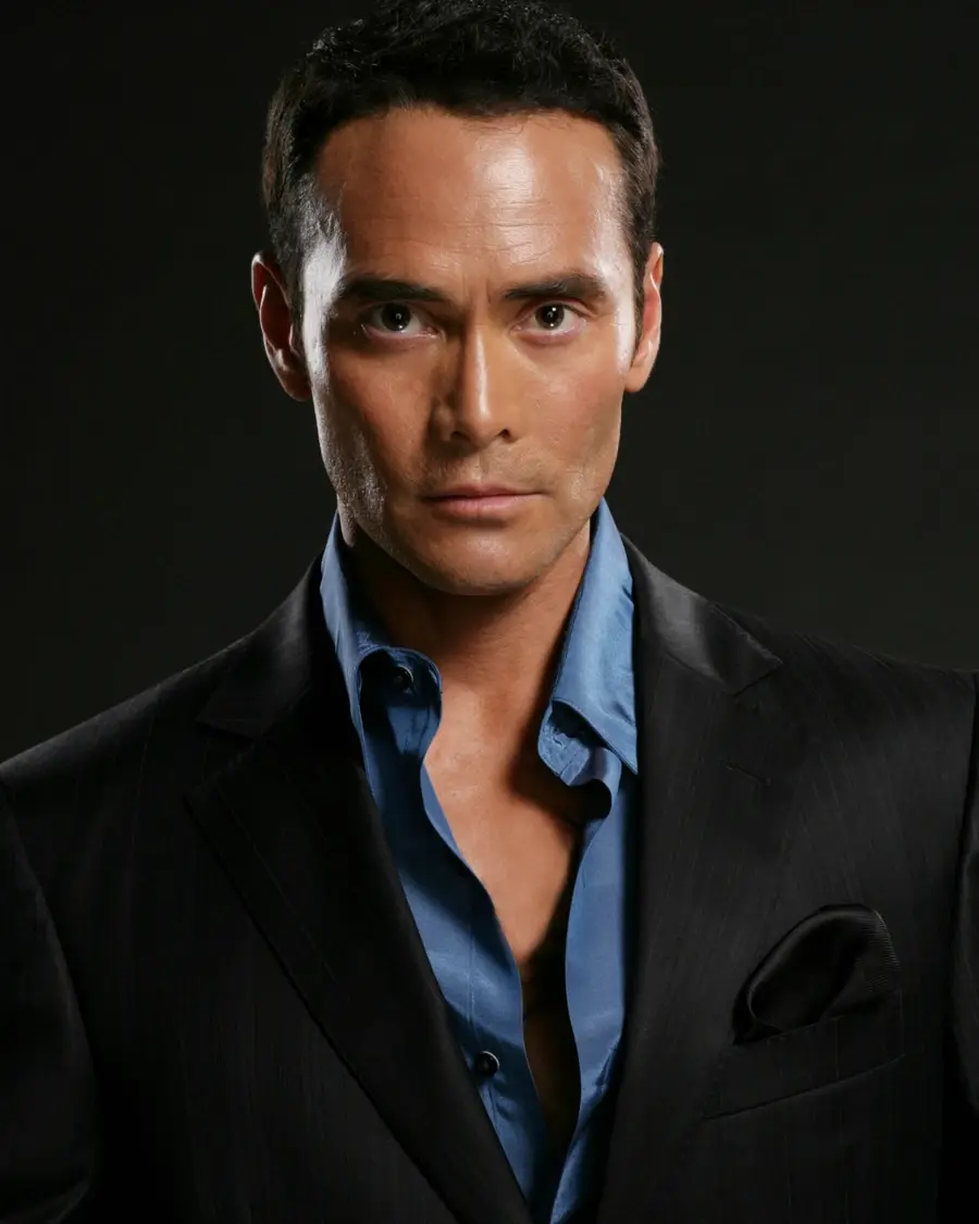 Man in a dark suit with a blue shirt looks intensely at the camera. Black background, formal attire, serious expression.
