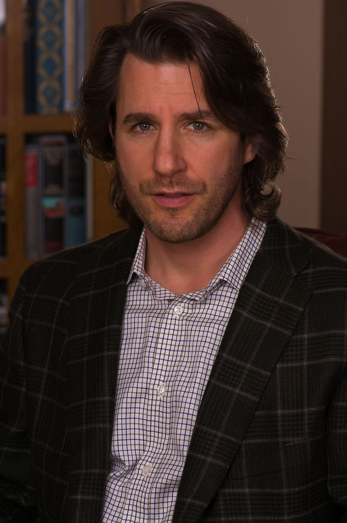 Man with long hair in a plaid blazer and checkered shirt, stands indoors against a bookshelf, appearing confident and relaxed.