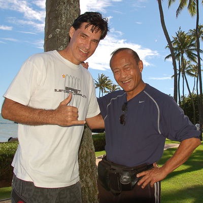 Two men smiling near a palm tree on a sunny beach, one wearing a white shirt and the other in a navy shirt. Bright sky and ocean in background.