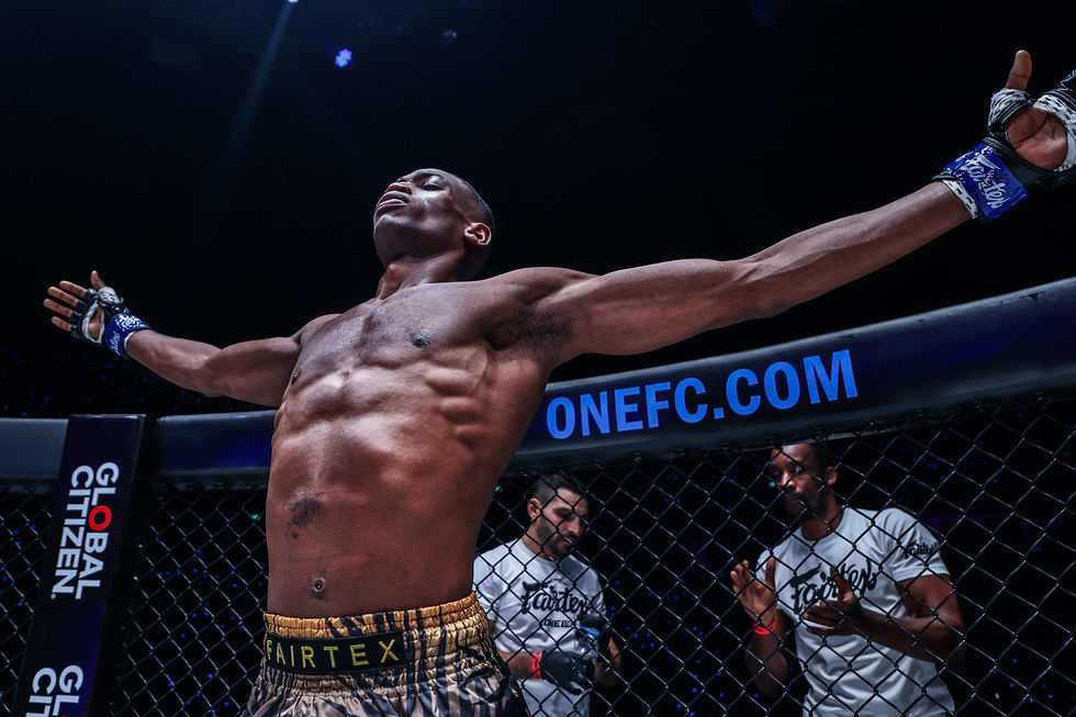 MMA fighter celebrates with arms spread in a cage, wearing gloves and shorts with "FAIRTEX." "ONEFC.COM" text on the cage. Emotive scene.