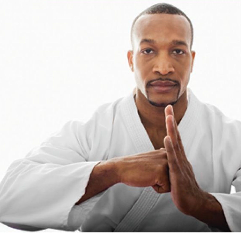 Man in a white karate gi performs a focused hand gesture, with a serious expression, against a plain white background.