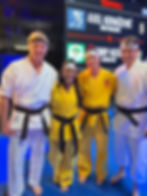 Four karate competitors in white and yellow gis smile on a blue mat before a Semifinals scoreboard showing Axel Kovačević vs Bobby Kee.