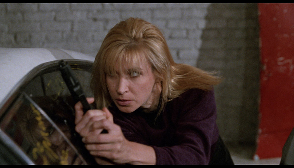 Blonde woman aiming a gun, crouched behind a car. Focused expression, brick wall background, red element visible on the side.