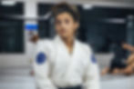 Woman in a white martial arts gi with blue patches sits in a gym, looking thoughtful. Two people grapple on the mat in the background.