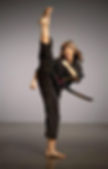 Woman in a black martial arts uniform performs a high kick with determination in a neutral studio setting. Her brown hair is flowing.