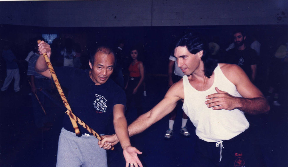 Two men practice Filipino martial arts with sticks in a dimly lit room. One wears a black shirt and gray pants, the other a white tank top.