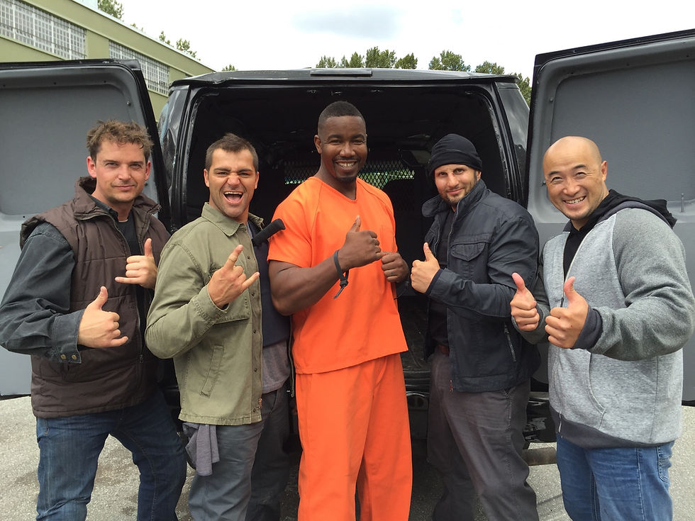 Five smiling men pose by an open black van, giving thumbs-up signs outdoors in front of a building.
