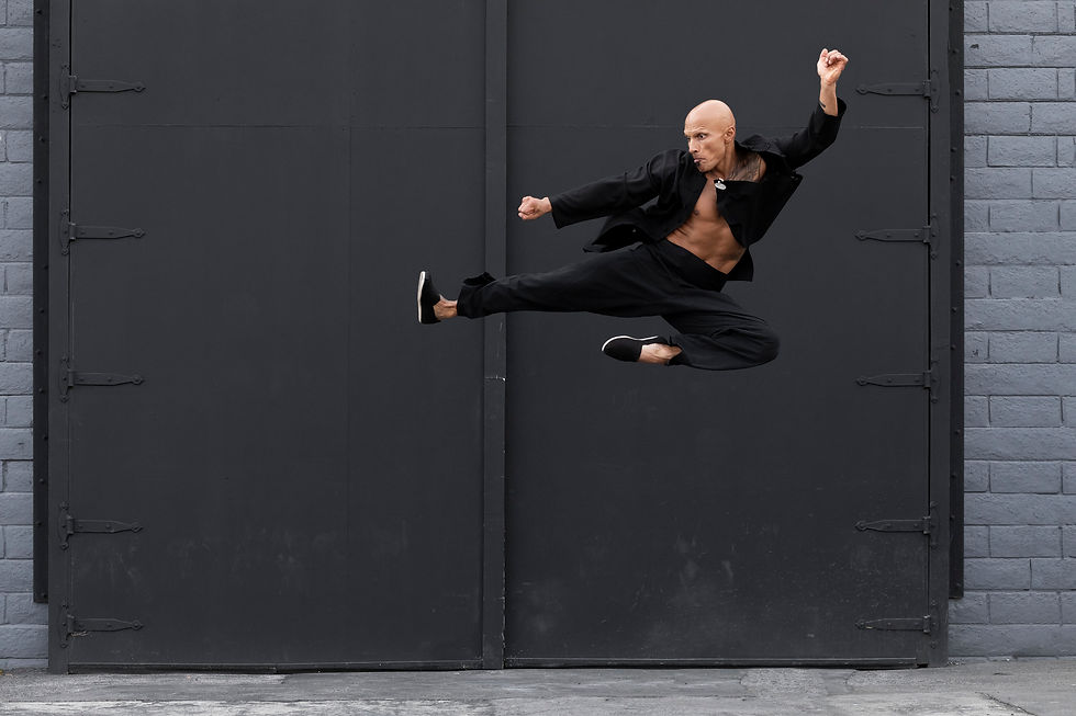 A bald man performs a dynamic martial arts jump kick against a gray industrial door backdrop, wearing all black attire.