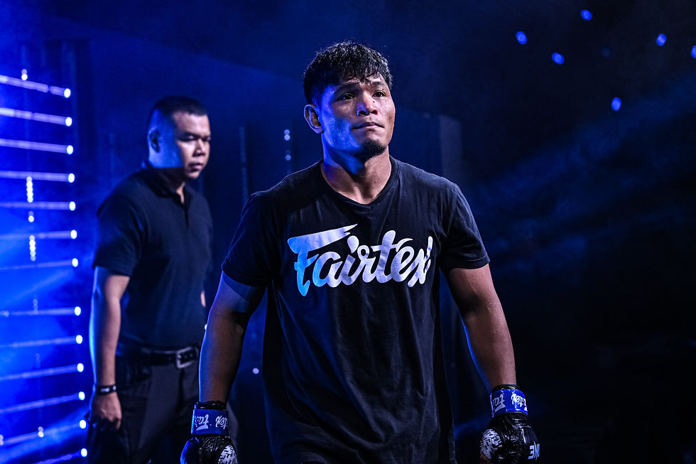 Man in black Fairtex shirt, blue gloves, walks purposefully in a dim, blue-lit setting; focused expression, another person in black nearby.