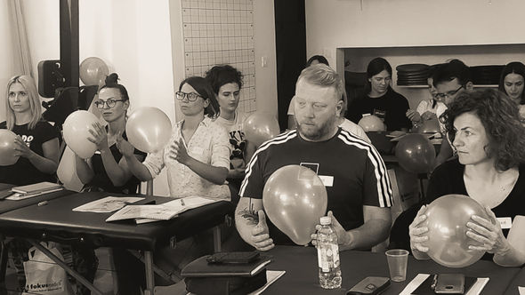 Crno-bela fotografija polaznika seminara manuelne tehnike miofascijalnog opuštanja u vežbi sa balonima u rukama, među terapeutskim stolovima u edukativnom centru.