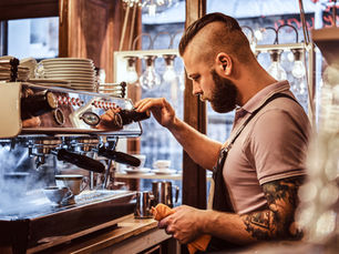 A tattooed barista in a cafe operates an espresso machine, concentrating as steam rises. Cups are stacked, ambient light glows softly.