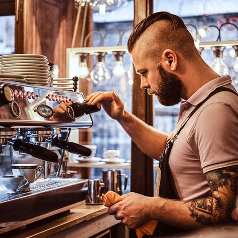 A tattooed barista in a cafe operates an espresso machine, concentrating as steam rises. Cups are stacked, ambient light glows softly.