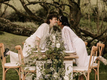 A female couple in wedding outfits leaning over a table for a kiss. A rustic table is styled with wild flowers, moss and vintage tableware - Dominic Williams Photography, Rudge Farm Weddings, Wild Soul Wedding Co Wedding Coordinator