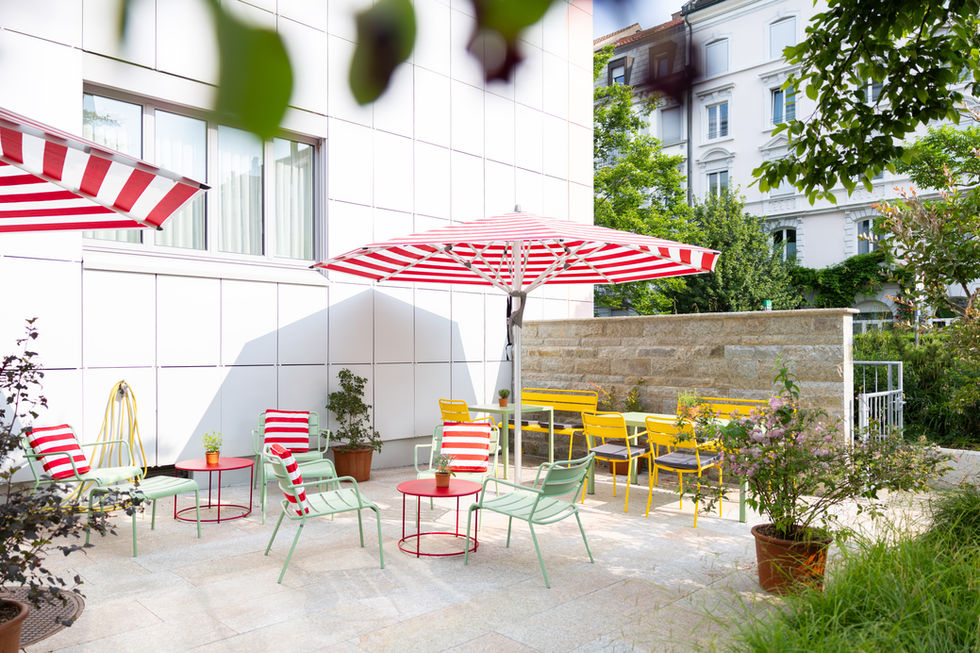 Pflegezentrum Falkenstein: Sitzterrasse, gelbe Sitzgarnitur mit Bank, mintgrüne Lounge Sessel mit rotem Clubtisch und rot weiss gestreiften Zierkissen, Sonnenschirm rot weiss gestreift, Topfpflanzen, Naturstein Wand und Boden, Farb- und Raumkonzept, Planung und Begleitung der Umsetzung: Patricia Leforestier, Basel, Schweiz