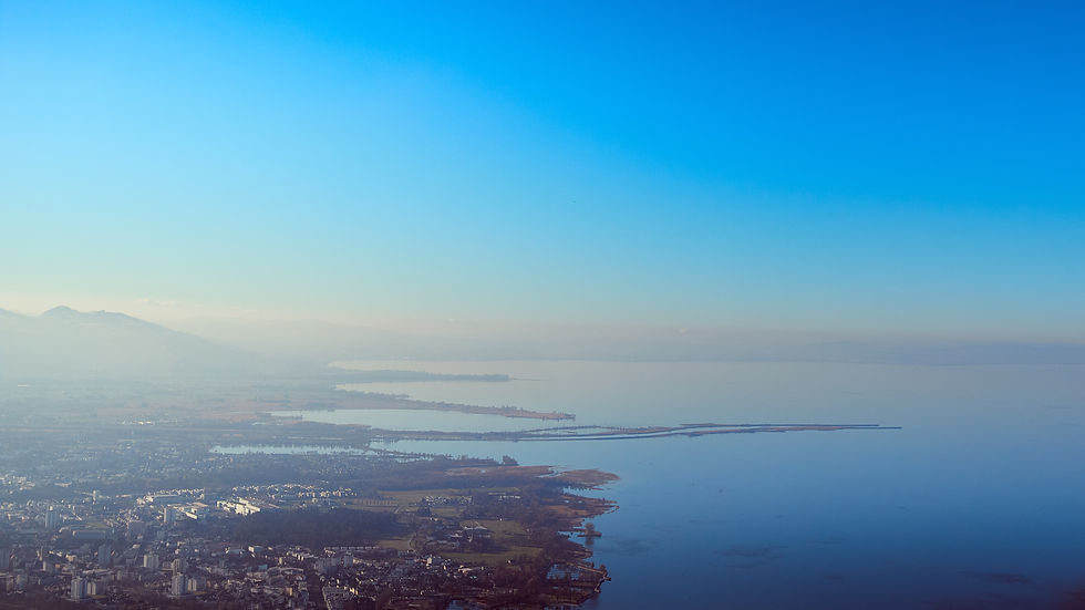 Luftaufnahme einer Stadt am See mit Dunst, Halbinseln und Bergen im Hintergrund unter blauem Himmel.